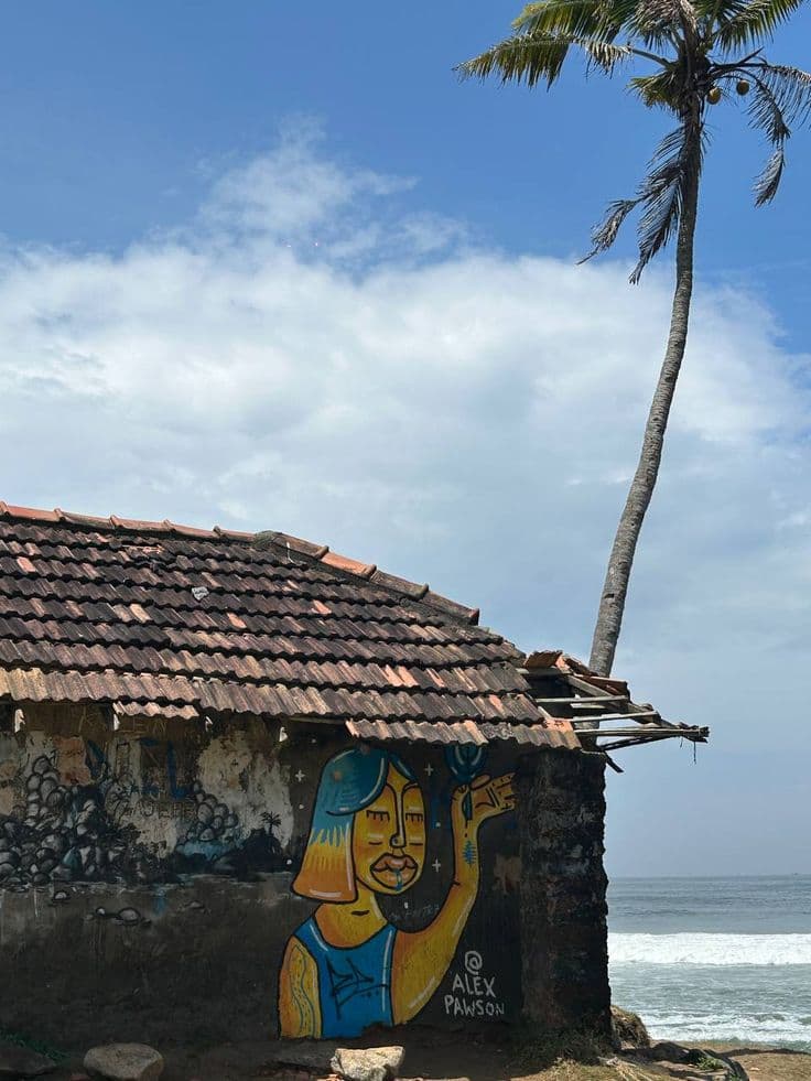 A tiled-roof house with a painted mural beside a palm tree and the sea